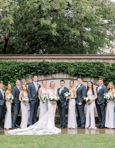 A wedding party poses outdoors, featuring the couple at the center surrounded by bridesmaids in light dresses and groomsmen in dark suits. Greenery and a stone wall form the background.